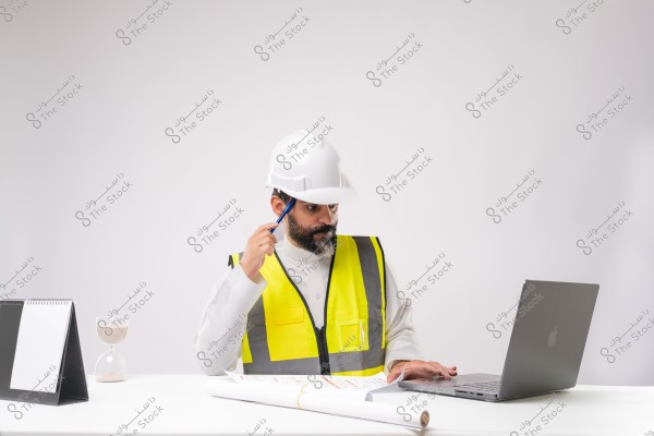 A photo of a man sitting at a desk wearing a white thobe, a high-visibility safety vest, and a white helmet. He appears to be an engineer or work supervisor working on a laptop. On the table in front of him are architectural blueprints, an hourglass, and a planner.