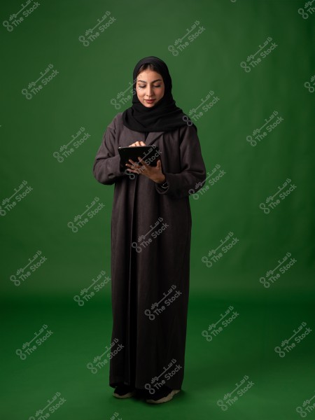 A portrait of a woman wearing a black abaya and hijab standing against a green background. She is holding a tablet and appears centered in the image. The lighting is bright, and the background is free from any prominent details or decorations.