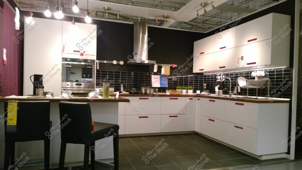 A modern kitchen with a corner design is displayed. It features white cabinets with red handles and a wooden countertop. A built-in silver oven and an extractor hood are above the stove. The backsplash is covered with black subway tiles and is cluttered with items like cups and small vases. Two black leather-backed chairs are in the foreground under the countertop.
