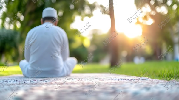 A man sitting on a prayer mat in a green park during sunset. He is wearing traditional white clothing with a cap and appears to be in a meditative or prayerful position, bathed in golden sunlight from the background, creating a serene and peaceful atmosphere.
