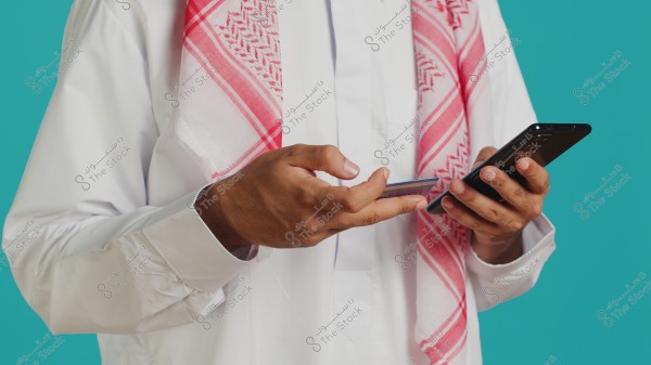The image shows a person wearing a traditional white thobe and a red checkered keffiyeh, holding a smartphone in the right hand and a card in the left hand. The background is blue.