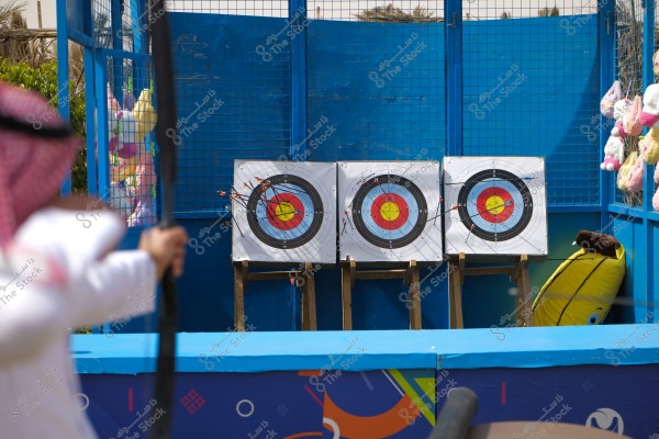 The image shows a person wearing a white thobe and a red and white checkered headscarf aiming a bow at three archery targets. The targets are colored and have multiple arrows protruding from different positions on the targets. The setting appears to be recreational or sporty, with a blue barrier and several stuffed toys visible in the background.\r\n\r\n###