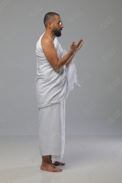 Image of a man wearing white Ihram clothing, standing in a prayer position. He is smiling and wearing sunglasses, with a neutral gray background.