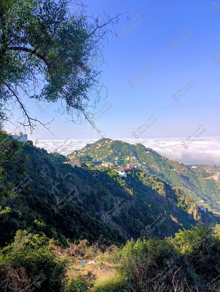 A natural landscape showing green mountains covered with trees and vegetation under a clear blue sky. In the background, some buildings are scattered on the mountain slopes with a part of the white clouds covering the horizon.