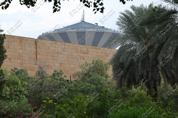 The image shows a palm tree and lush garden in front of a sandy-colored stone wall. Dominating the center of the image is a large dome- or saucer-shaped tower in gray and white, topped with an antenna. The foreground is filled with green trees and plants, while the sky appears slightly cloudy in the background.