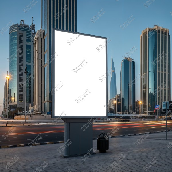 A large blank billboard stands in the center of a modern urban scene. Tall skyscrapers surround the billboard in the background, suggesting the location might be in a Gulf city like Dubai. The road shows light traffic with light trails from passing cars, and the sky is clear blue, indicating it\'s daytime.