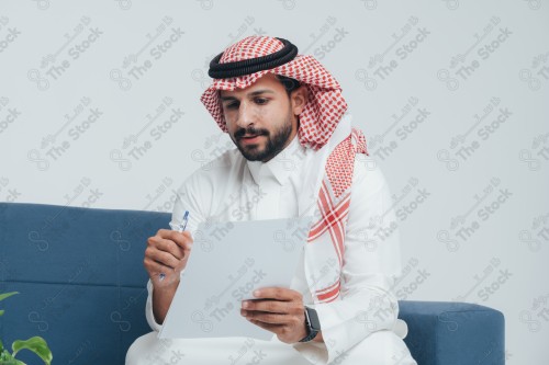 رجل سعودي يرتدي الزي السعودي التقليدي يجلس على أريكة ويراجع ورقة عمل