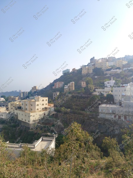 A scenic view of a mountainous city with a collection of light-colored buildings constructed on slopes. Trees and vegetation fill the areas surrounding the buildings, and the sky is clear blue, providing a sense of peace and calm. The image highlights the mountainous terrain and the natural environment of the area.