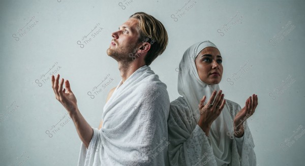 Image of a man and a woman wearing traditional white attire, standing side by side, with their hands raised in a prayer position. The man is dressed in a white cloth, and the woman wears a white hijab adorned with intricate patterns. The background is plain white.