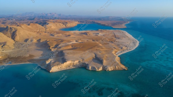 Aerial view of a desert coastline running alongside a blue sea. The image highlights a striking contrast between the yellow sands of the desert areas and the clear blue waters of the sea. Mountainous desert terrain is visible in the background with a clear sky on the horizon.