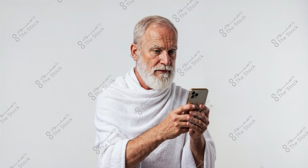 A portrait of an elderly man with a white beard and gray hair, wearing a white robe resembling an ihram garment. He is holding a smartphone and looking at it intently. The background is plain white.