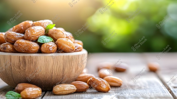 The image shows a wooden bowl filled with glossy dates, aesthetically arranged with some small green leaves for decoration. Dates are also scattered on a wooden surface in the foreground, and the background features a blurred green hue, evoking a natural scene with early morning or sunset lighting.