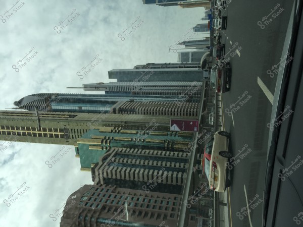 The image shows towering skyscrapers in a modern city, with cars driving on a highway. The buildings feature glass and modern architecture, and the sky appears cloudy. There are signs indicating the presence of shops and road signs visible in the image.