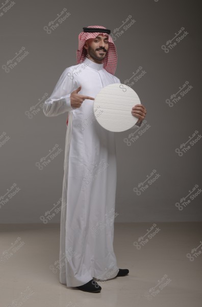 A portrait of a man standing in a studio wearing traditional Saudi attire, a white thobe, and a red checked headscarf (shemagh) with a black headband (agal). The man is smiling and holding a white circular object, pointing at it with his hand. The background is plain gray.