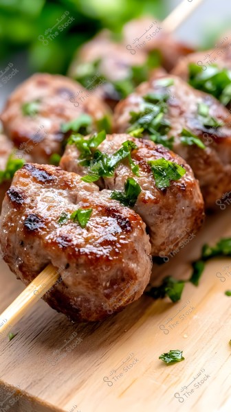 Image of grilled meatballs skewered on wooden sticks. The meatballs are perfectly grilled with char marks and a sheen indicating how they were cooked. The top of the meatballs is garnished with chopped green herbs, adding a touch of freshness and color. The background contains more green leaves, giving the image depth.