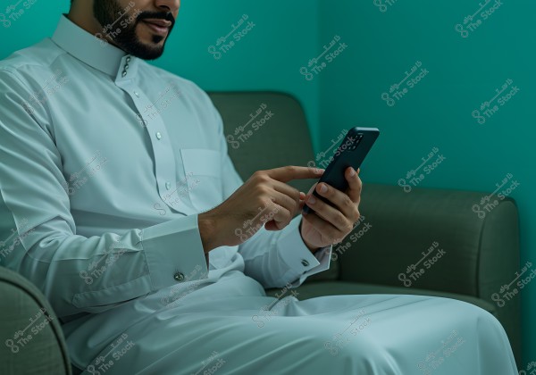 The image shows a man sitting on a chair, wearing a white thobe, and using a mobile phone. The wall and the background are turquoise. This image might be from Saudi Arabia, given the traditional thobe.