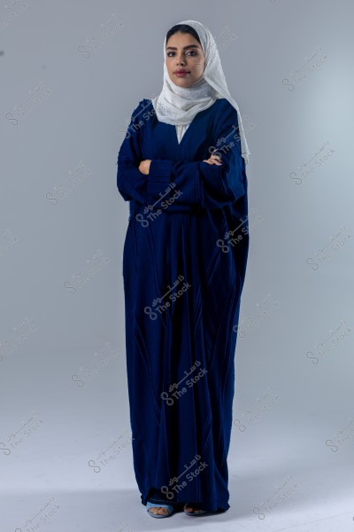 An image of a woman wearing a dark blue abaya and a white headscarf. She stands with her arms crossed, looking confidently at the camera. The background is neutral, highlighting the details of her clothing.