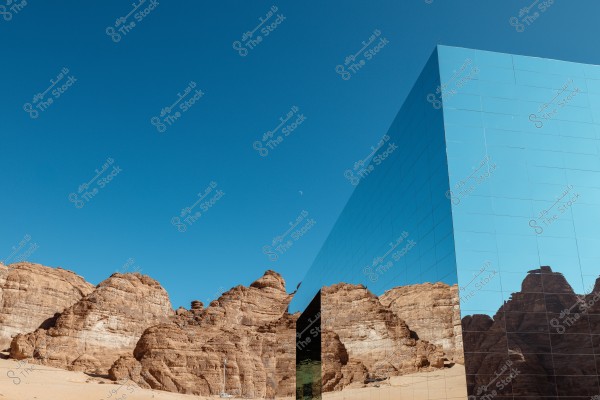An image depicting a desert landscape under a clear blue sky. On the right, there is a modern building with a mirrored facade reflecting the surrounding large desert rock formations, creating a unique visual balance between nature and architecture.