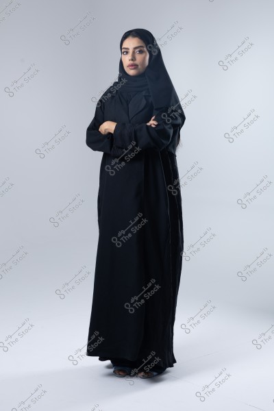 Portrait of a woman standing against a light gray background, wearing a black abaya and a black hijab. She has a serious expression with her arms crossed in front of her.