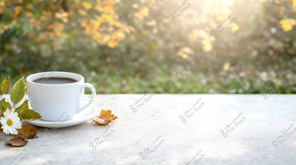 A cup of coffee in a white cup on a white table in sunlight. Next to the cup, there is a white flower and some green and golden leaves. The background features greenery and autumn leaves in brown and gold colors.
