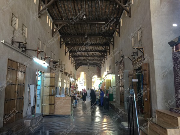An image of an indoor passage in a traditional market featuring a wooden ceiling with iron beams. On either side of the passage, there are shops with wooden doors displaying various goods. The floor is tiled with traditional patterns. A few people in traditional clothing are visible in the background, walking through the passage.