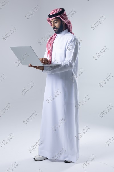 An image of a man wearing a white thobe and a red and white ghutra, working on a laptop. He stands against a white background, looking intently at the screen. The clothing suggests that it is from Saudi Arabia.