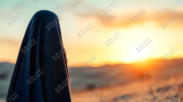 The image shows a side view of a person covered in a dark cloak standing against a sunset. The sky is filled with shades of orange and gold, and the surrounding landscape features soft hills and thin clouds on the horizon. The image evokes a sense of calmness and mystery.