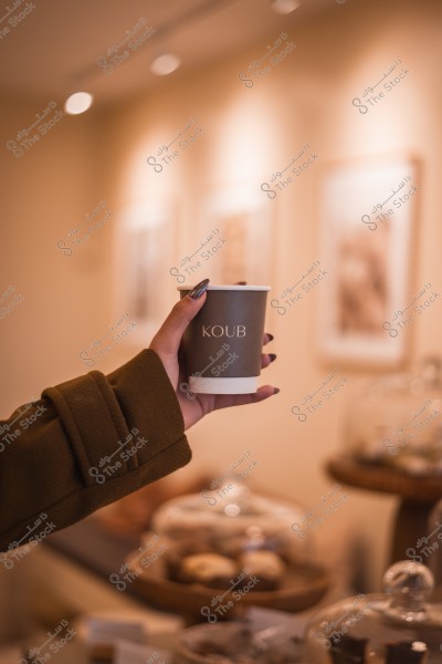 A hand holding a coffee cup labeled \"KOUB\" in a warmly lit café with pastries displayed in the background.