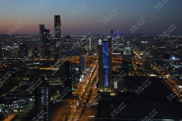 A night view of a large city with vibrant lighting and a skyline illuminated by towering buildings. A tall building in the foreground is lit with distinctive blue lights, surrounded by a cluster of modern structures and busy roads glowing with night lights. The city appears to sparkle under a dark sky with a gradient of sunset colors.