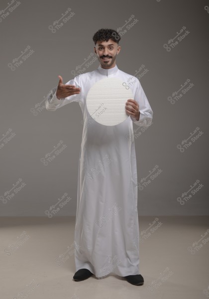 An image of a man wearing a traditional white robe, standing against a grey background. He is holding a large white circle in his right hand, while his left hand is raised pointing at the circle. His expression appears happy or cheerful.