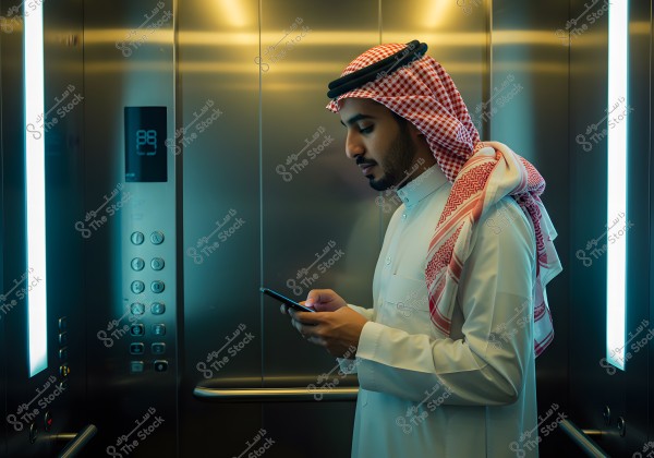 A portrait of a man wearing traditional attire and a shemagh standing in a modern elevator. The elevator appears to be made of metal, giving it a sleek look. The man is focused on his mobile phone, appearing engaged. The elevator\'s lighting is balanced, creating a soft atmosphere. Elevator buttons and the number panel are visible on the left side of the image.