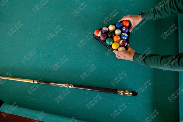 The image shows a green pool table with a person arranging billiard balls in a triangle. The balls are of various colors such as red, yellow, and blue. A pool cue is placed on the table, and the person\'s arms are in long-sleeved green clothing.
