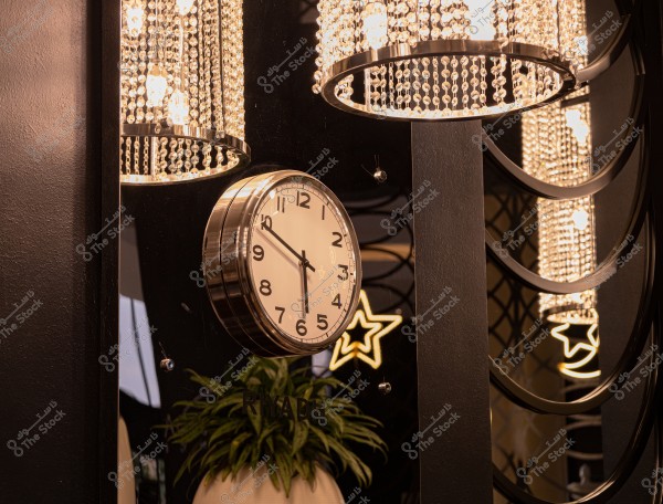 تُظهر الصورة ساعة حائط بتصميم كلاسيكي وبإطار معدني مرآوي, مثبتة على جدار داكن. تعكس الساعة بريق أضواء من ثريات مزينة بأعمدات بلورية. تحت الساعة, يظهر نبات في إناء أبيض. يوجد في الخلفية تصميم على شكل نجمة مضيئة, مما يضفي على المكان جواً فخماً.