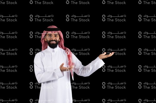 بورتريه رجل سعودي يرتدي الثوب، يقف على خلفية سوداء ويقوم بعمل حركات بيديه لعرض او وصف منتج، رجل يقوم بعمل حركات بيديه وهو ينظر الى الكاميرا