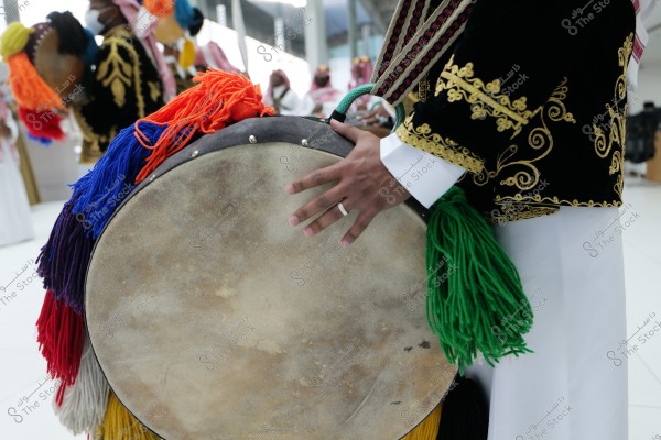 An image shows a person wearing traditional attire with gold embroidery holding a large drum adorned with multicolored fabric tassels, including red, blue, and green. In the background, other individuals in similar cultural clothing are engaged in a celebration of local heritage, indicating a cultural or festive event. The setting appears to be a modern, well-lit hall.