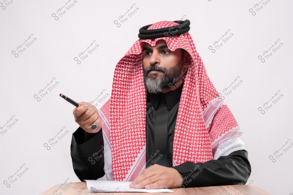 A man wearing a black thobe and red ghutra, seated at a table, holding a pen and a paper.