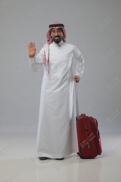 An image of a man wearing a traditional Saudi white thobe, a red and white checkered ghutra, and a black agal, standing and smiling while holding a red suitcase. He is waving with his hand. The background is grey.