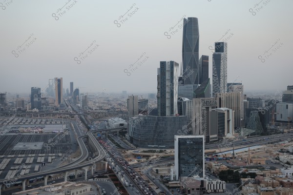 Aerial view of Riyadh, Saudi Arabia, showcasing a collection of modern skyscrapers and distinctive architectural designs. The King Abdullah Financial District building stands out prominently, with a network of highways in the foreground. Surrounding urban areas and residential buildings spread out under a cloudy sky.