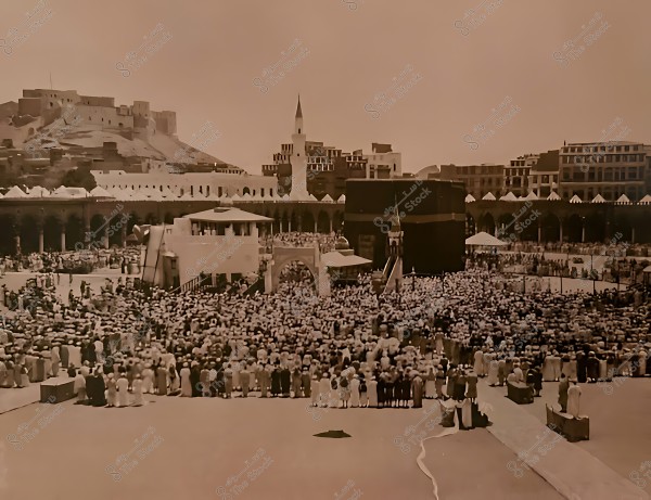 A historical photo of the Kaaba in Mecca with crowds of people in traditional attire gathered around it. The Grand Mosque is visible in the background with its minarets and old buildings.