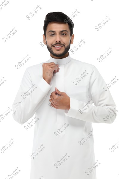A portrait of a man wearing a white thobe, which is a traditional garment often associated with Saudi Arabia. The man is smiling and adjusting the collar of his thobe, appearing ready for the photo. The background is white.