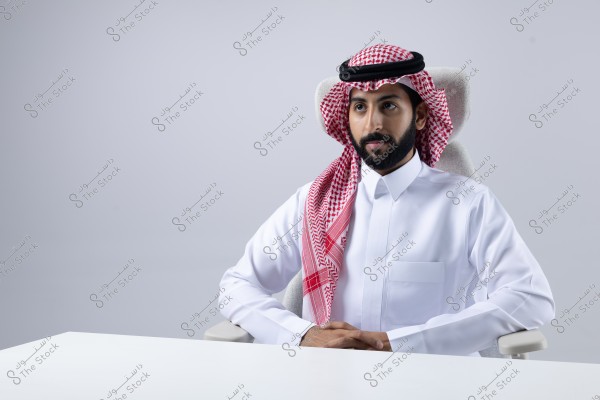 A portrait of a man sitting on a chair wearing traditional Saudi attire, a white thobe, and a red shemagh with a black agal. The man is seated at a white table with a light gray background.