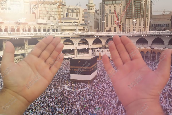 Hands raised in prayer in front of the Kaaba at the Grand Mosque in Mecca, with crowds of pilgrims.