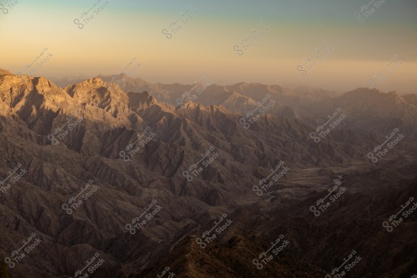 A scenic view of a vast wavy mountain range with prominent rocky peaks in the foreground. The mountain is bathed in golden sunlight at sunset, casting warm shadows across the textured terrain. The sky is clear blue above, transitioning to a bright orange near the horizon.