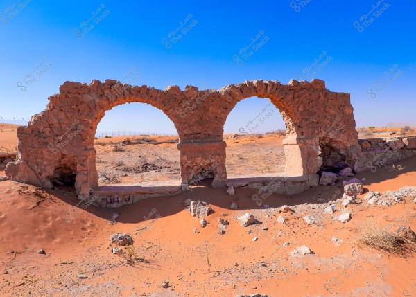 The image shows the remnants of a stone structure with three arches located in a desert with orange sand. The background features a clear blue sky, and there are some dry grass and small scattered rocks on the ground.