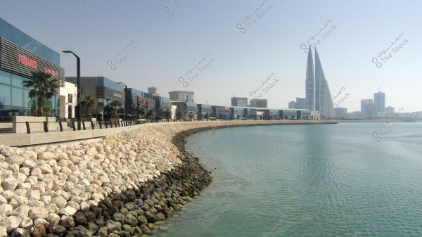 Image of a waterfront in Manama, Bahrain, showing a row of shops and restaurants with brand signs on modern reflective glass buildings. There is a promenade by the sea lined with large stones. In the background, the \"Bahrain World Trade Center\" towers are visible with their distinctive triangular glass design.