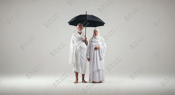 Image of a man and a woman standing side by side against a white background. The man is holding a black umbrella and wearing a white Ihram, while the woman is dressed in a white hijab and robe. Their attire reflects the appearance of Muslim pilgrims performing Umrah or Hajj.