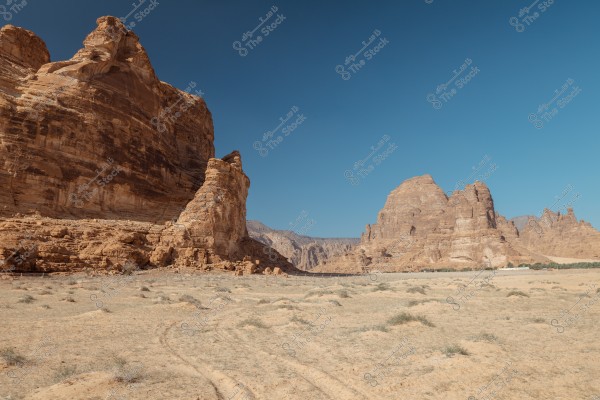 A natural landscape of a desert featuring large majestic rock formations under a clear sky. The rocks are a dark brown color with distinct lines and terrains, and the ground is covered with sand and sparse desert vegetation.