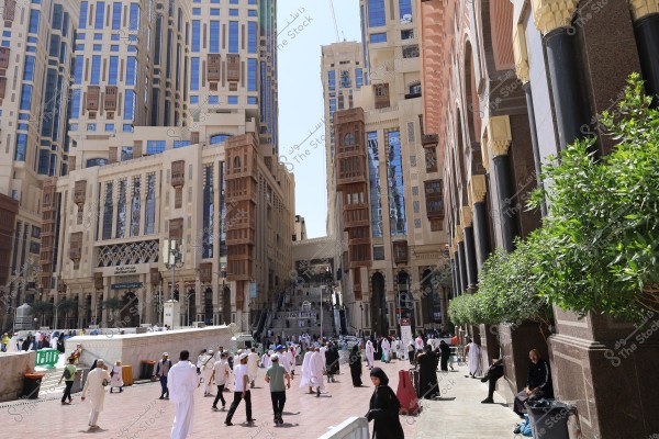 The image shows a busy street in Mecca, Saudi Arabia, with a crowd of people wearing traditional Saudi attire, such as white thobes, ghutras, and agals, while women wear abayas. The surrounding high-rise buildings feature modern Islamic architecture combined with traditional elements like lattice wooden windows.