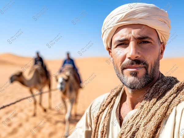 Image of a man wearing a white turban and traditional clothing in the desert. In the background, a camel is being led by two people also in traditional attire. The sky is clear blue, and the golden sand stretches in all directions.