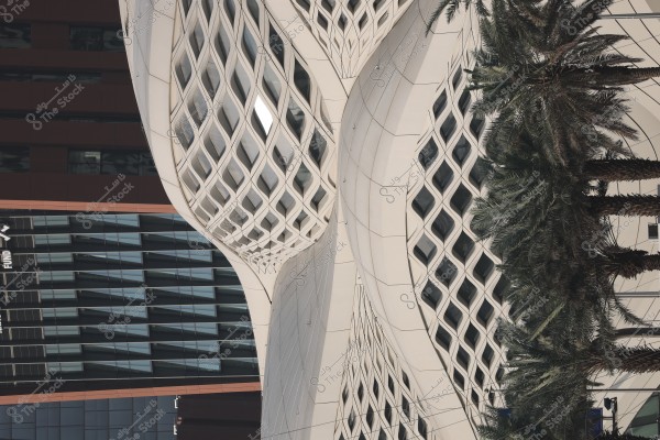 Image of a building facade with a modern and intricate architectural design, featuring white lattice patterns on a glass exterior. Palm trees are visible to the side, adding a natural element to the urban scene.